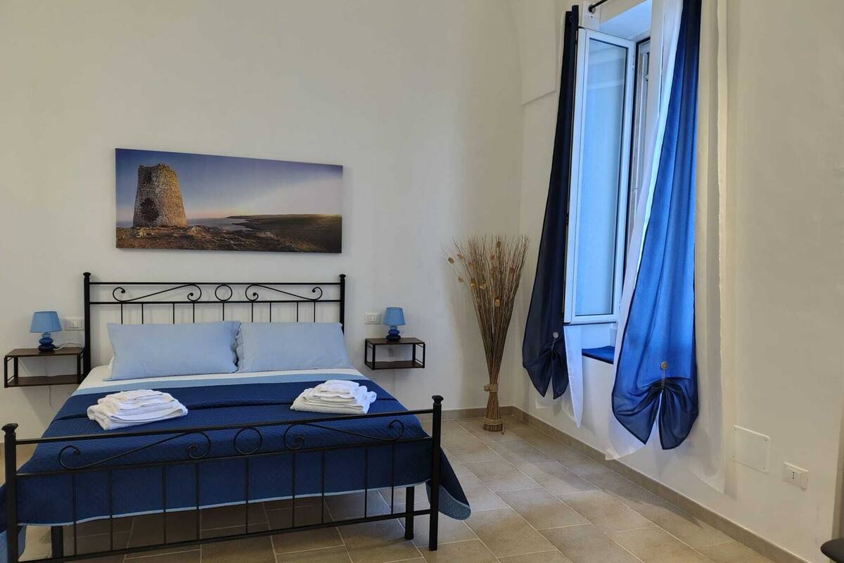 Un dormitorio con una cama, dos mesitas de noche, una lámpara de noche, una ventana con cortinas azules, un cuadro en la pared y un florero con plantas secas.