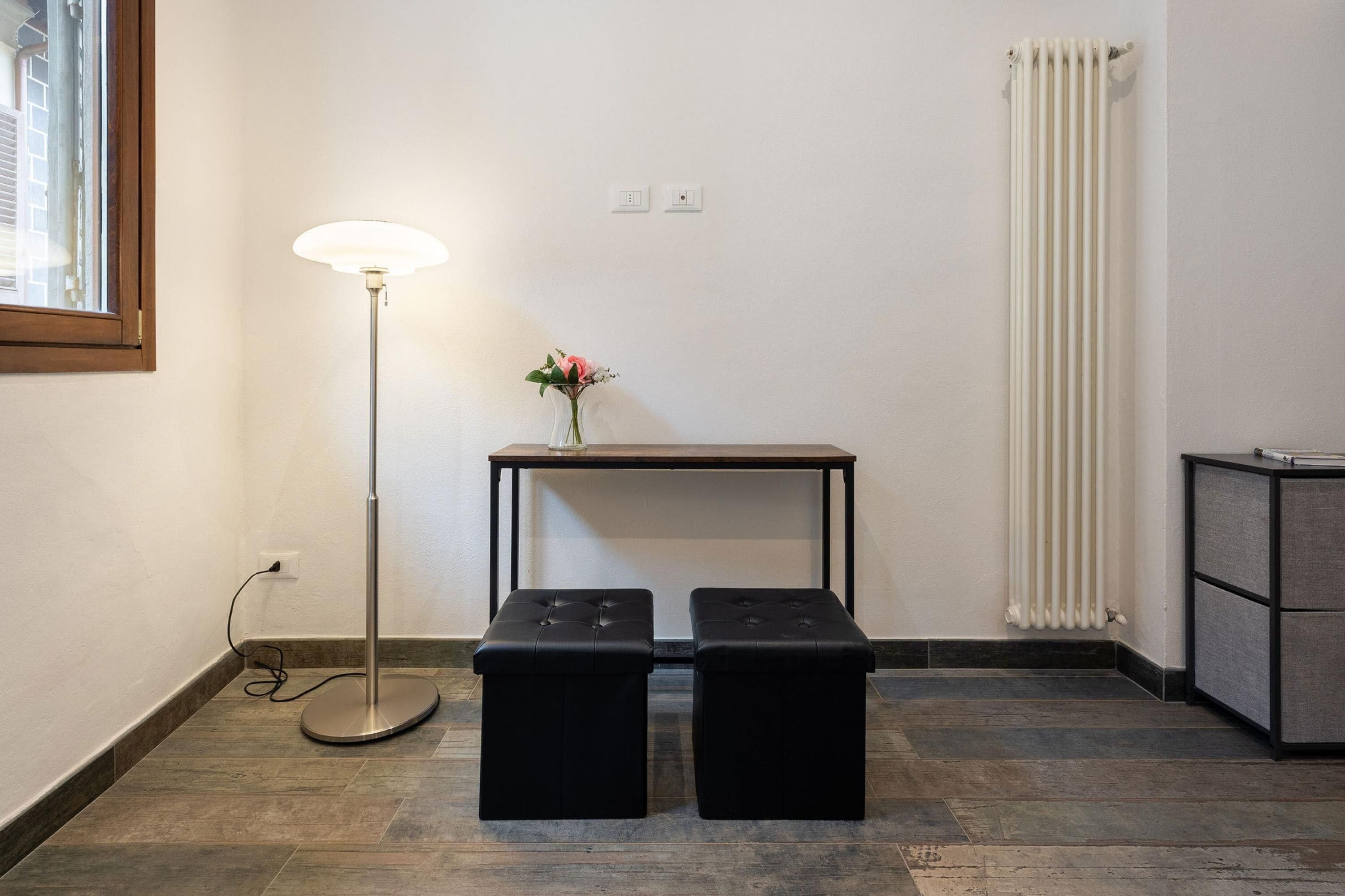 Ein Zimmer mit einer stehenden Stehlampe, einem kleinen Tisch mit Blumenvase, zwei schwarzen Hocker, einem Heizkörper und einem Schrank.