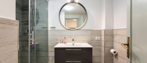Un bany amb dutxa de vidre, un mirall rodó, un lavabo blanc amb un moble fosc i un vàter.