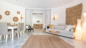 A modern living room with a white sofa, a dining table with chairs, and a coffee table. There are wall-mounted lights and a woven wall hanging. - Pool Villa Rancho Domingo Family Friendly (Benalmádena)
