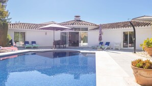 Una piscina con sillas de descanso y sombrillas, rodeada por un edificio con techo de tejas y puertas de cristal.