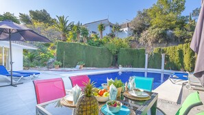 Un área de comedor junto a la piscina con una mesa preparada para dos personas, rodeada de sillas coloridas, una mesa de vidrio y una piscina al fondo.