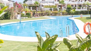A swimming pool with a clear blue water, surrounded by greenery and a white building in the background. - Sunset Golf Groundfloor Shared Pool (Estepona)