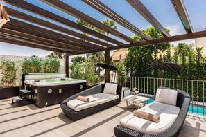 Una terraza en la azotea con un jacuzzi, dos sillones de descanso, una mesita pequeña y una piscina al fondo.