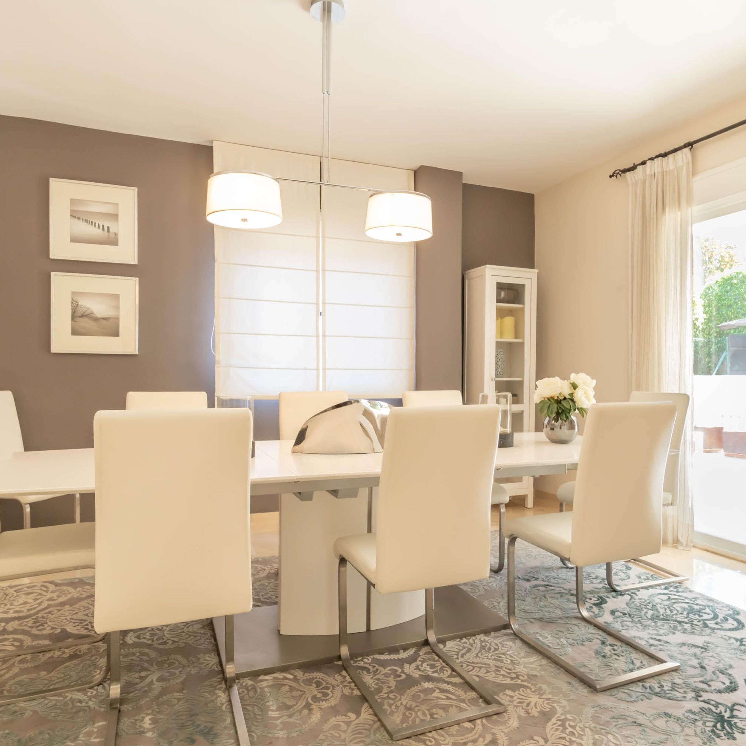 Un comedor moderno con una mesa blanca, cuatro sillas y una araña. Hay cuadros en la pared y un aparador con un jarrón de flores.