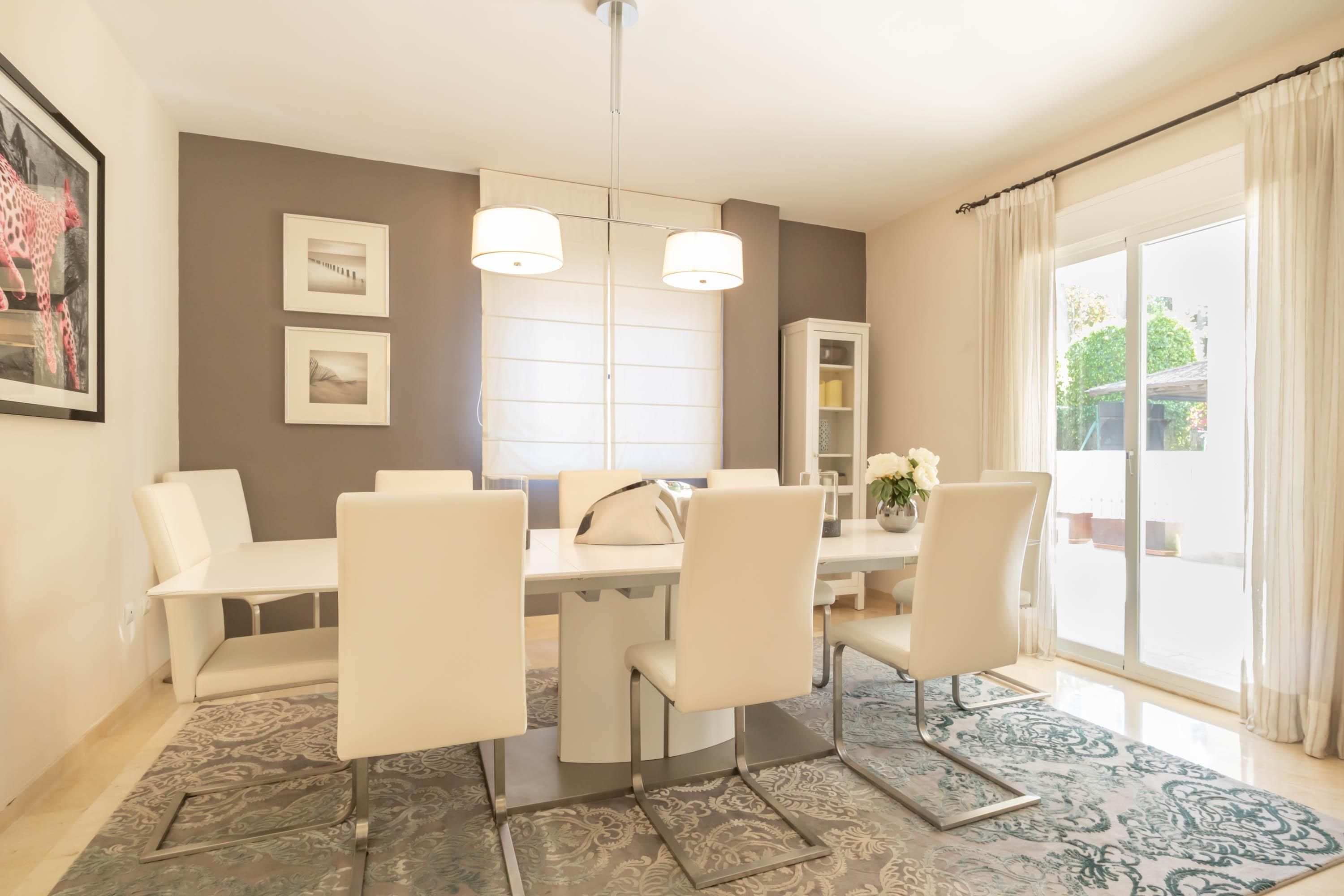 Un coin repas avec une table et des chaises blanches, un lustre et un tapis à motifs.