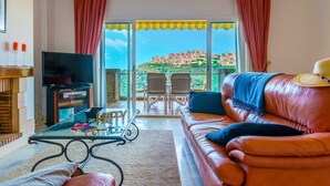En stue med en lædersofa, et glasbord, et fladskærms-tv og en balkon med udsigt til bygninger.