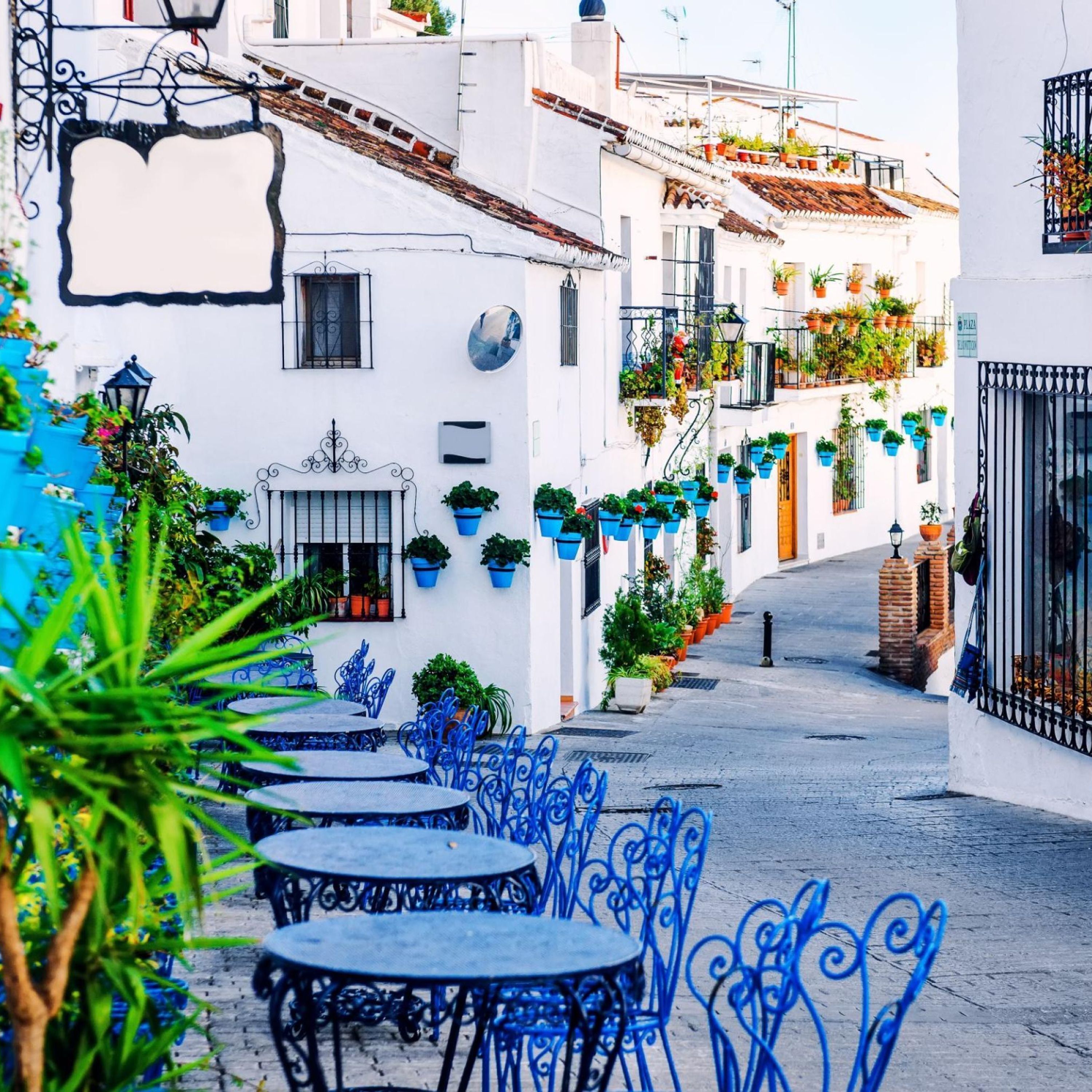A narrow street with white buildings, blue outdoor seating, and potted plants.