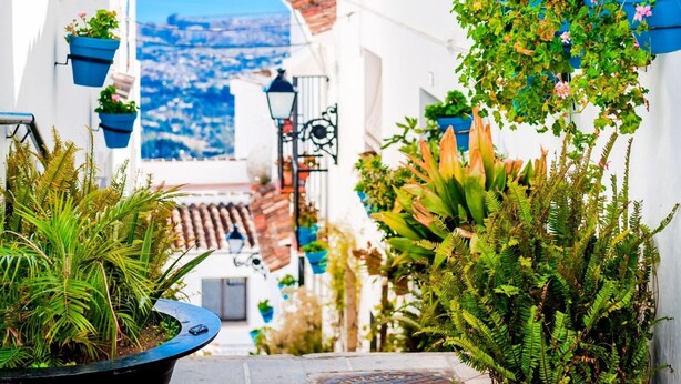 Una stretta via fiancheggiata da edifici bianchi, con fioriere blu appese e vista sul mare in lontananza.