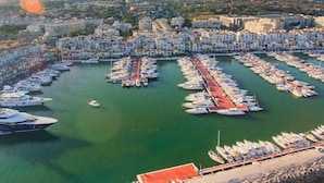 Μια αεροφωτογραφία μιας μαρίνας, όπου πολλά σκάφη είναι αγκυροβολημένα σε μια περιοχή, διατεταγμένα σε ένα γεωμετρικό σχέμα, ενώ γύρω τους υπάρχουν οικιστικά κτίρια και στο βάθος ορίζονται βουνά.