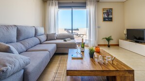 A modern living room with a grey sofa, a wooden coffee table with a bottle and glasses, a TV on a white cabinet, and a balcony with a view. - Sunset Seaview Corner With Pool (Estepona)