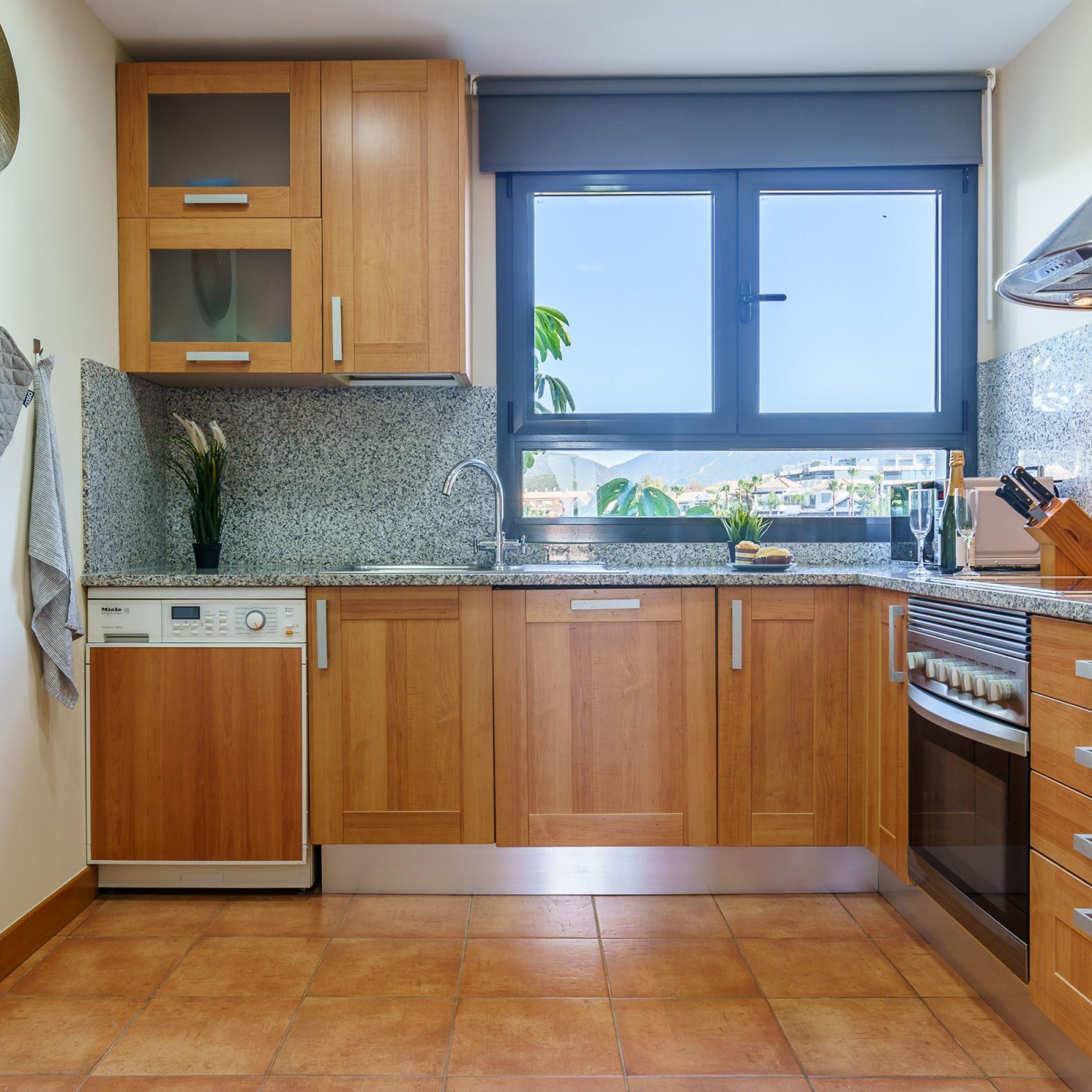 A kitchen with wooden cabinets, a built-in dishwasher, a window with a view, and a countertop with a knife block and wine glasses.