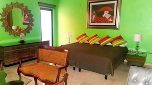 Habitación de hotel con pared verde, cama con almohadas de colores, una silla de madera, escritorio con espejo y un cuadro enmarcado en la pared.