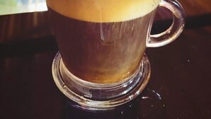 Una taza de vidrio transparente con una bebida en capas, posiblemente café, sobre una superficie reflectante.