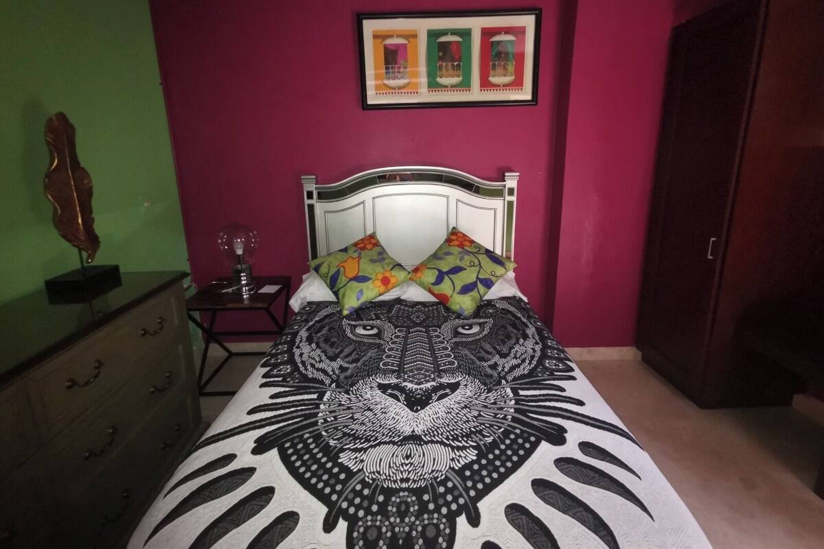 Un dormitorio con una cama que presenta un diseño de rostro de león en blanco y negro, un aparador de madera, una mesita de noche con una lámpara y un cuadro en la pared.