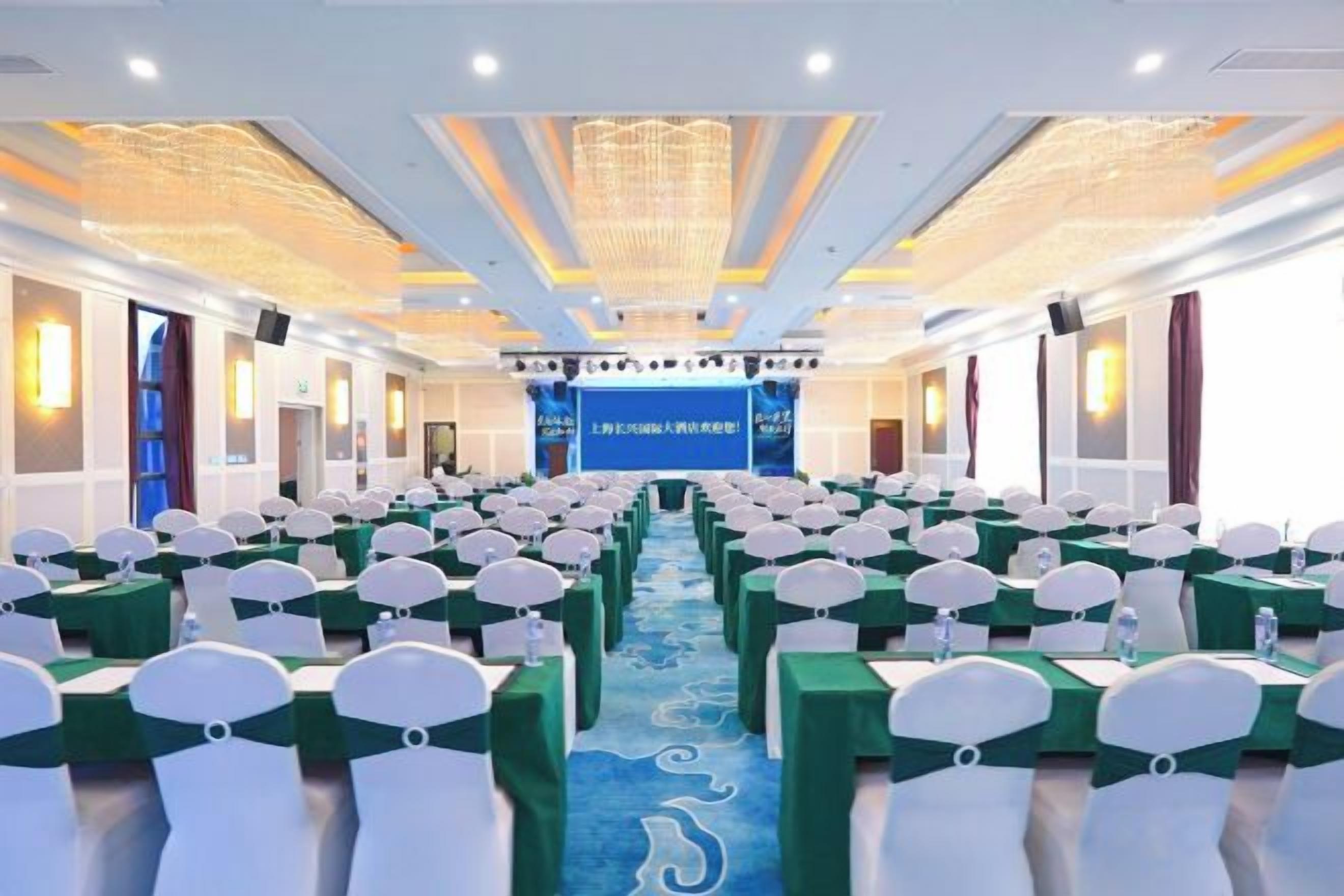 A conference room with rows of tables and chairs, a stage with a blue backdrop, and chandeliers.