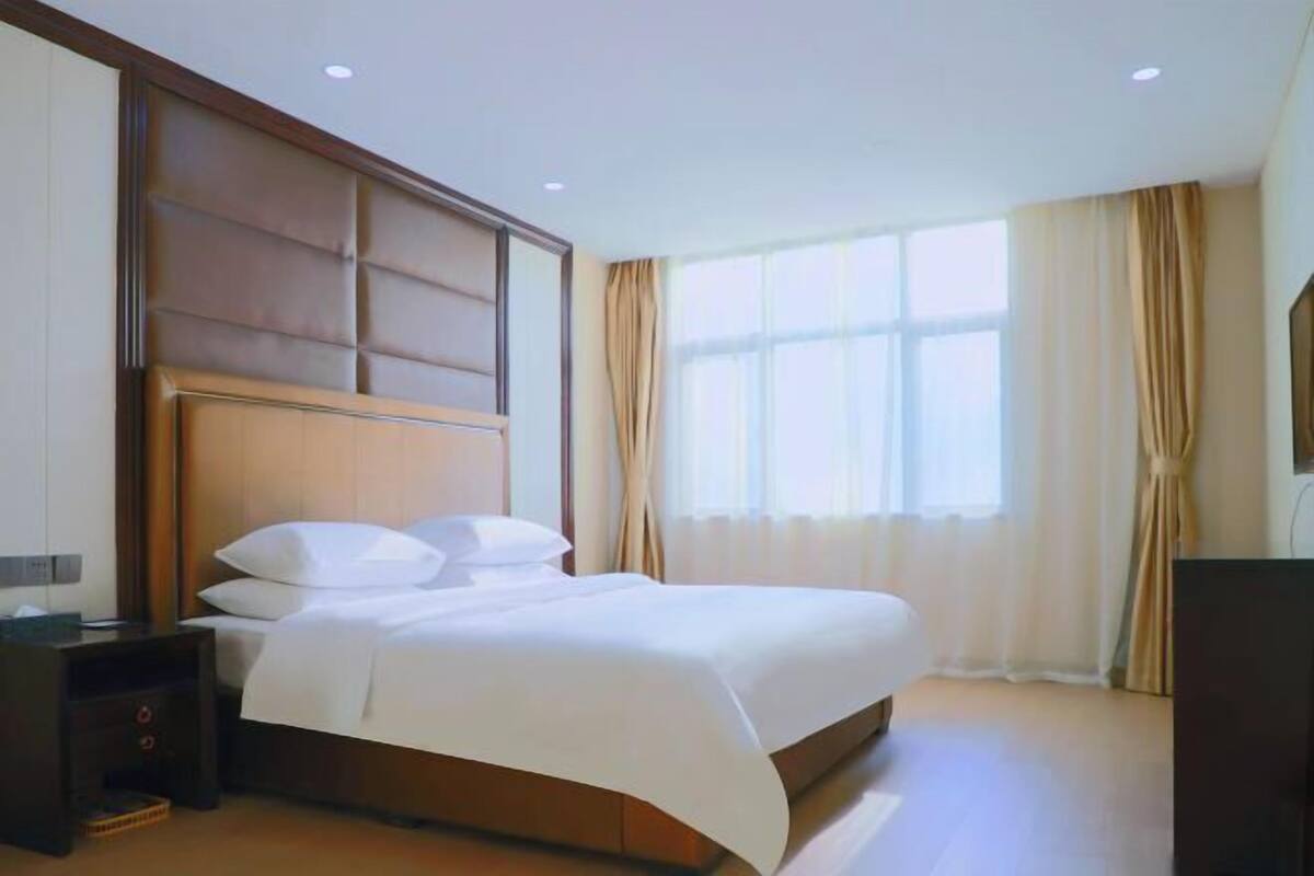 A hotel room with a large bed, a wooden headboard, a bedside table, a TV, and a window with curtains.
