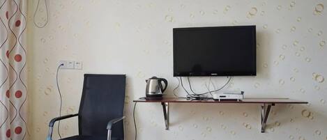 Habitación con televisión de pantalla plana montada en la pared, una silla, una mesita con hervidor y un papel tapiz estampado.