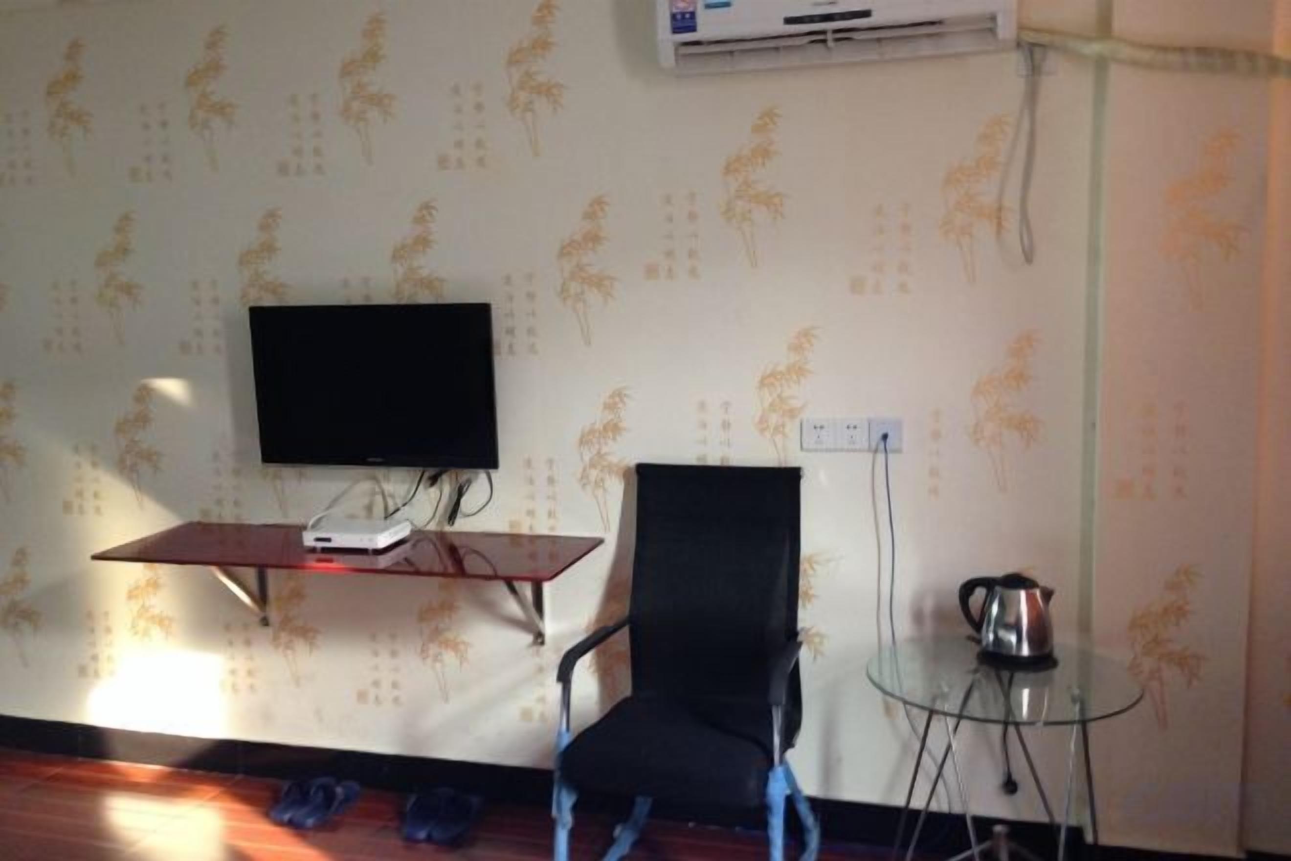 Una habitación con televisión de pantalla plana sobre un soporte, una silla negra, una mesita de cristal con un hervidor y papel tapiz estampado.