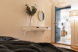 Ein Schlafzimmer mit einem Bett, einem Nachttisch mit Spiegel, einer Vase mit Grünpflanzen und einer Tür zu einem anderen Raum.