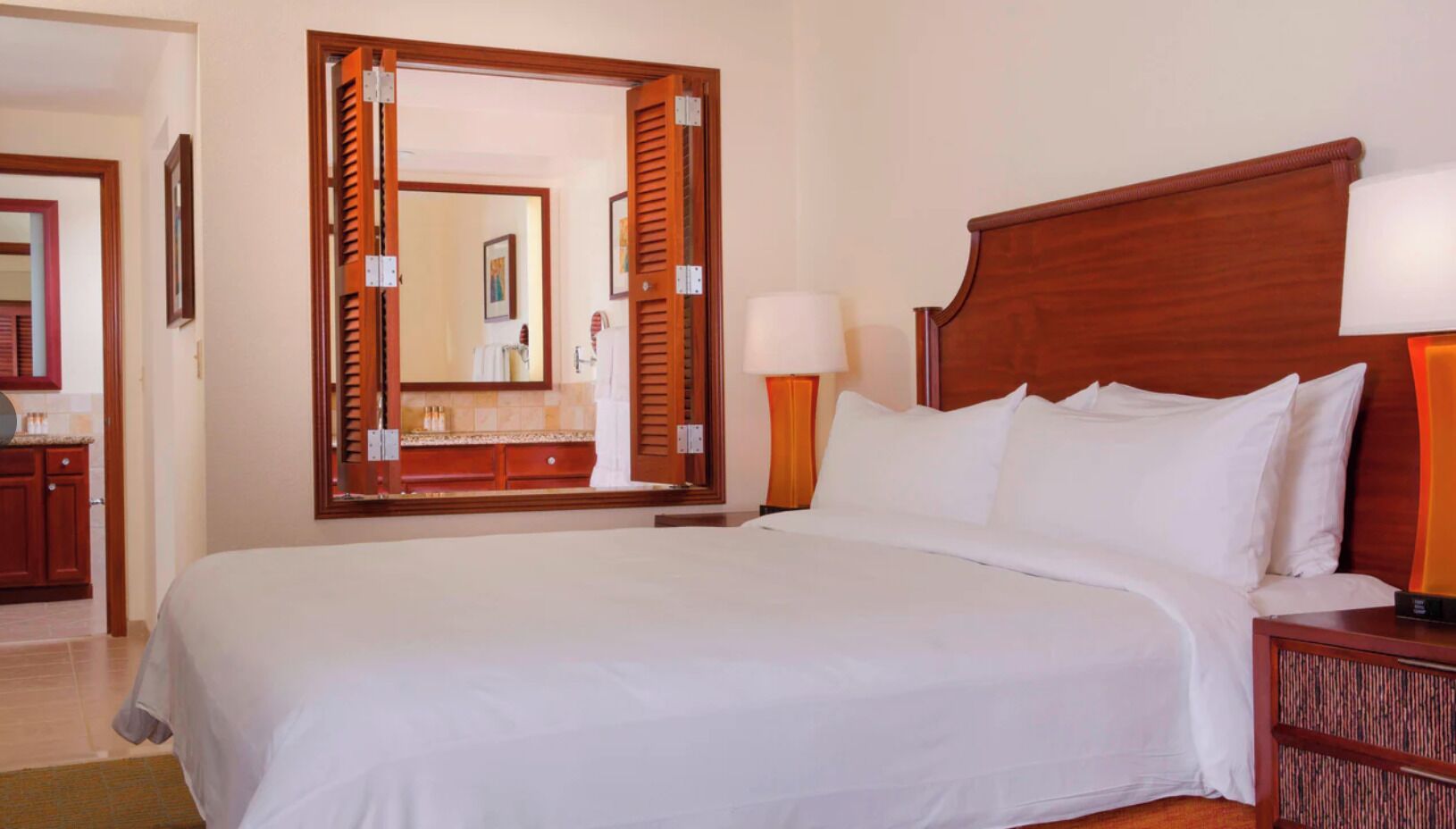 Een hotelkamer met een groot bed, een houten hoofdbord, een nachtkastje met lamp en een spiegel die de badkamer weerspiegelt.