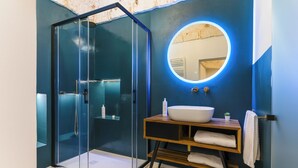 Un baño moderno con ducha acristalada, un espejo redondo con luz y un tocador de madera con lavamanos y toallas.