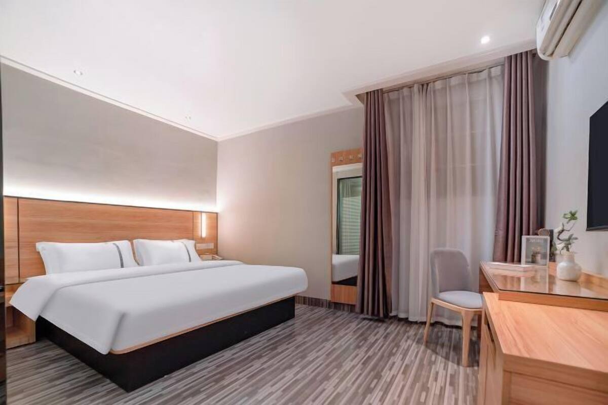 A hotel room with a bed, a desk, a chair, a mirror, and an air conditioning unit.