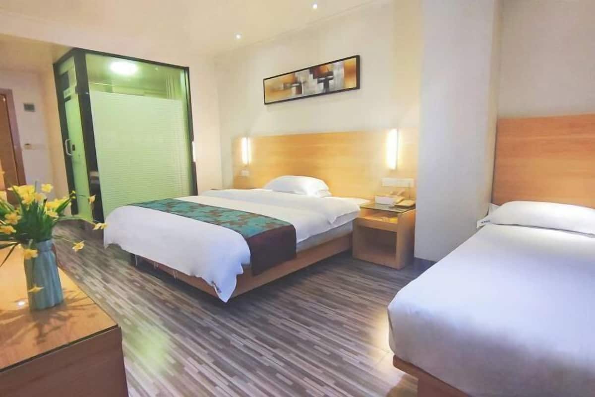 A hotel room with a large bed, a bedside table, and a nightstand. There is a window with blinds, a painting on the wall, and a vase with flowers on the nightstand.