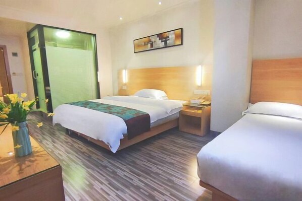 A hotel room with a large bed, a bedside table, and a nightstand. There is a window with blinds, a painting on the wall, and a vase with flowers on the nightstand. - Huajing Hotel(Yulin Miaoyuan) (Yulin)
