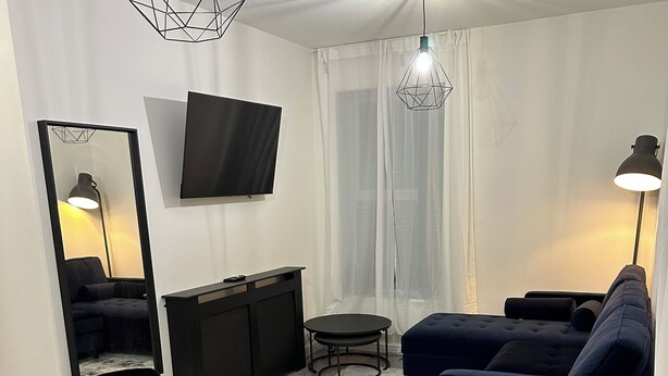 A modern living room with a dining table, black chairs, a blue sofa, a TV mounted on the wall, and a large mirror.