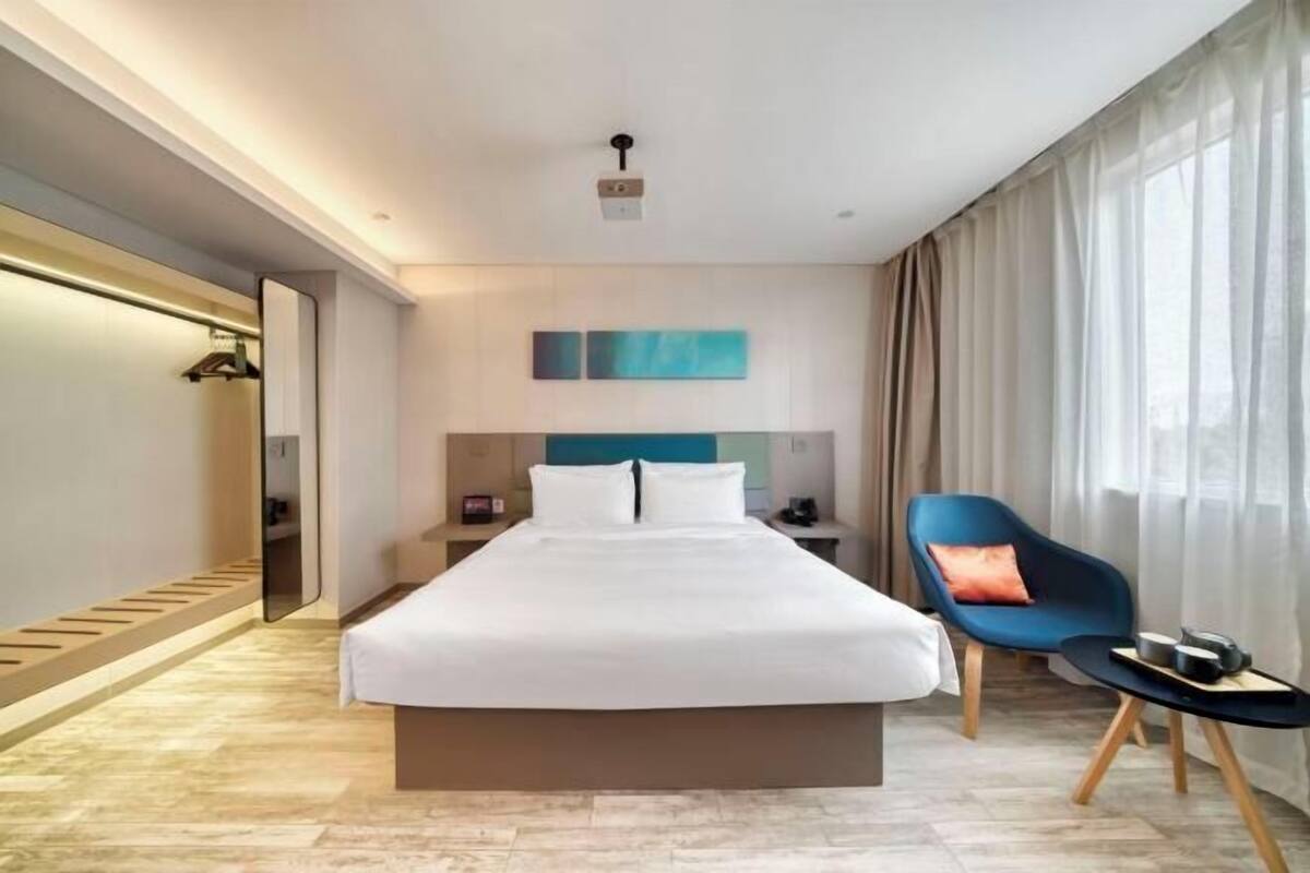 Una camera d'albergo moderna con un grande letto, un comodino, una poltrona blu e vista sull'esterno.