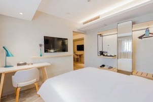 Quarto de hotel com cama, escrivaninha, cadeira, televisão e espelho.