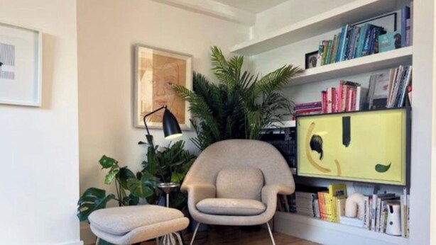 Une pièce d’angle avec un plancher en bois, un fauteuil rond, un petit tabouret, une bibliothèque et une grande plante en pot.