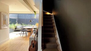 Un escalier menant à un étage supérieur sombre, un coin repas avec une table ronde et des chaises, et une guitare appuyée contre un pilier.