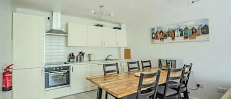 A modern kitchen with white cabinets, a wooden dining table, and a painting of colorful houses on the wall.