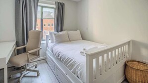 Un dormitorio con cama, escritorio, silla, ventana con cortinas y una cesta de mimbre.