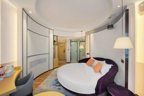 Une chambre d’hôtel moderne avec un lit aux lignes courbes, un bureau avec une chaise, une lampe et une vue sur le couloir.