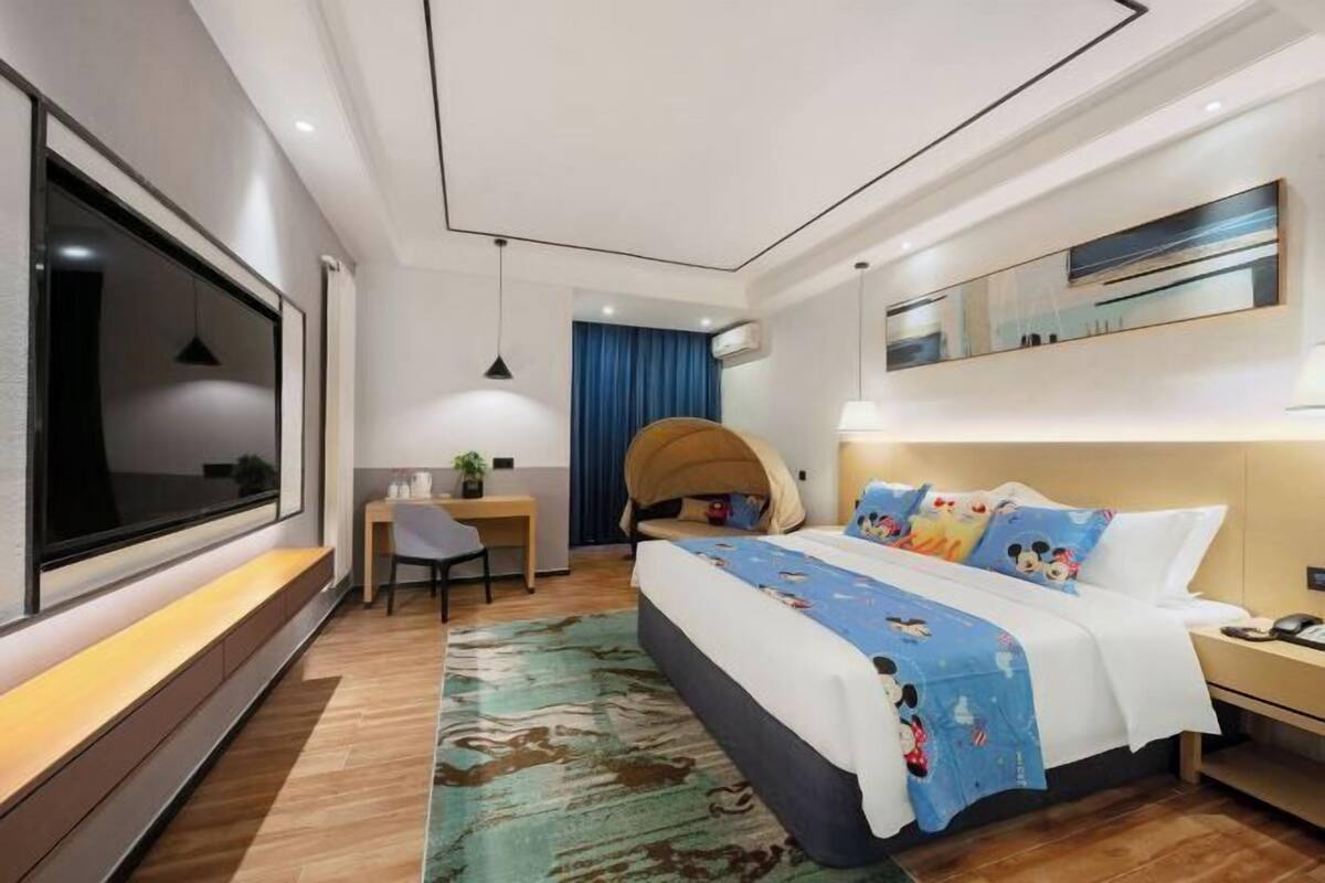 A modern hotel room with a large bed, a desk, a chair, a TV, and a large window.