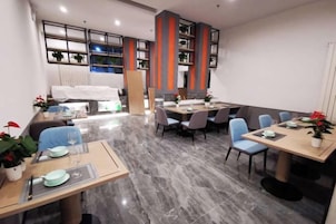 Un comedor moderno de hotel con varias mesas y sillas, una cocina al fondo y vistas de la ciudad a través de amplios ventanales.