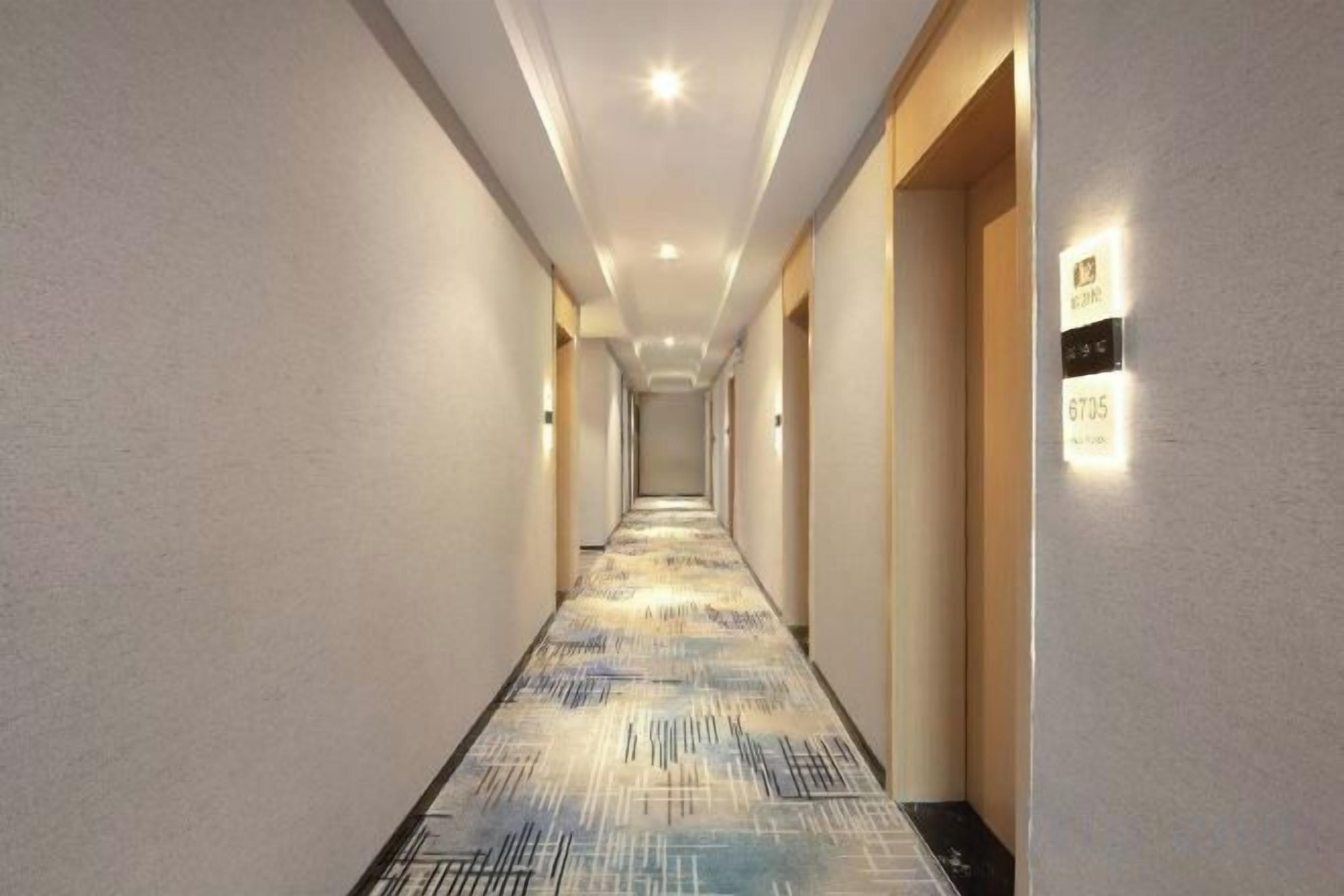 A hotel hallway with a patterned carpet, wall-mounted light fixtures, and a door with a number plate.