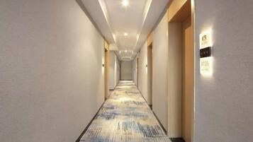 A hotel hallway with a patterned carpet, wall-mounted light fixtures, and a door with a number plate.