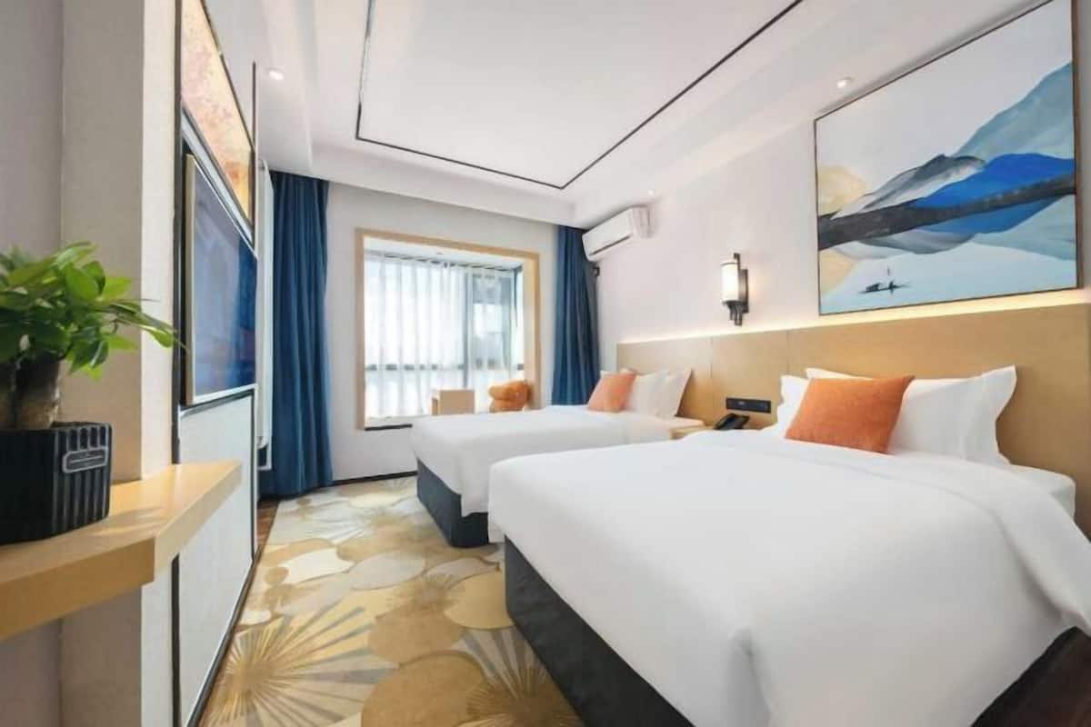 A hotel room with two beds, a large window with curtains, a plant on the side table, and a painting on the wall.