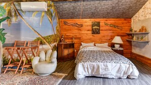 A bedroom with a wooden ceiling, a bed with a patterned bedspread, a white armchair, a wooden nightstand, and a wall mural of palm trees.