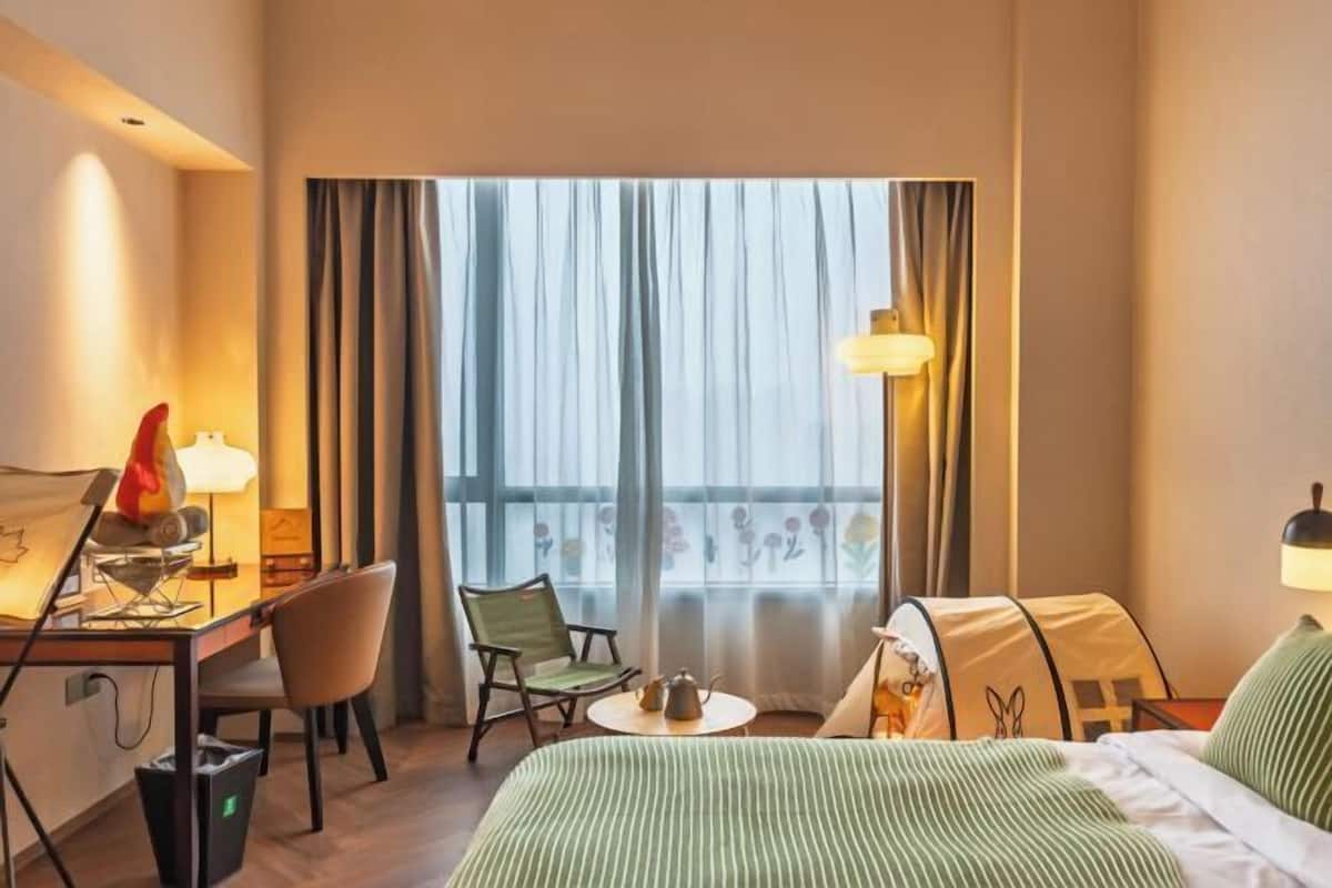 Ein Hotelzimmer mit einem Bett, einem Schreibtisch mit Lampe, einem Sessel, einem kleinen Tisch mit Teekanne und Blick ins Freie durch große Vorhänge.