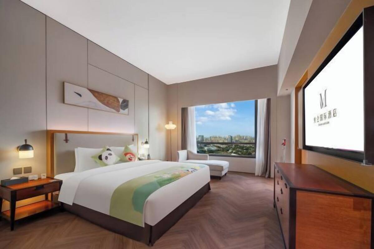 Ein Hotelzimmer mit Bett, Nachttischen, einem Schreibtisch, einem Sessel und einem großen Fenster mit Blick auf die Stadt, sowie einem an der Wand befestigten Fernseher.