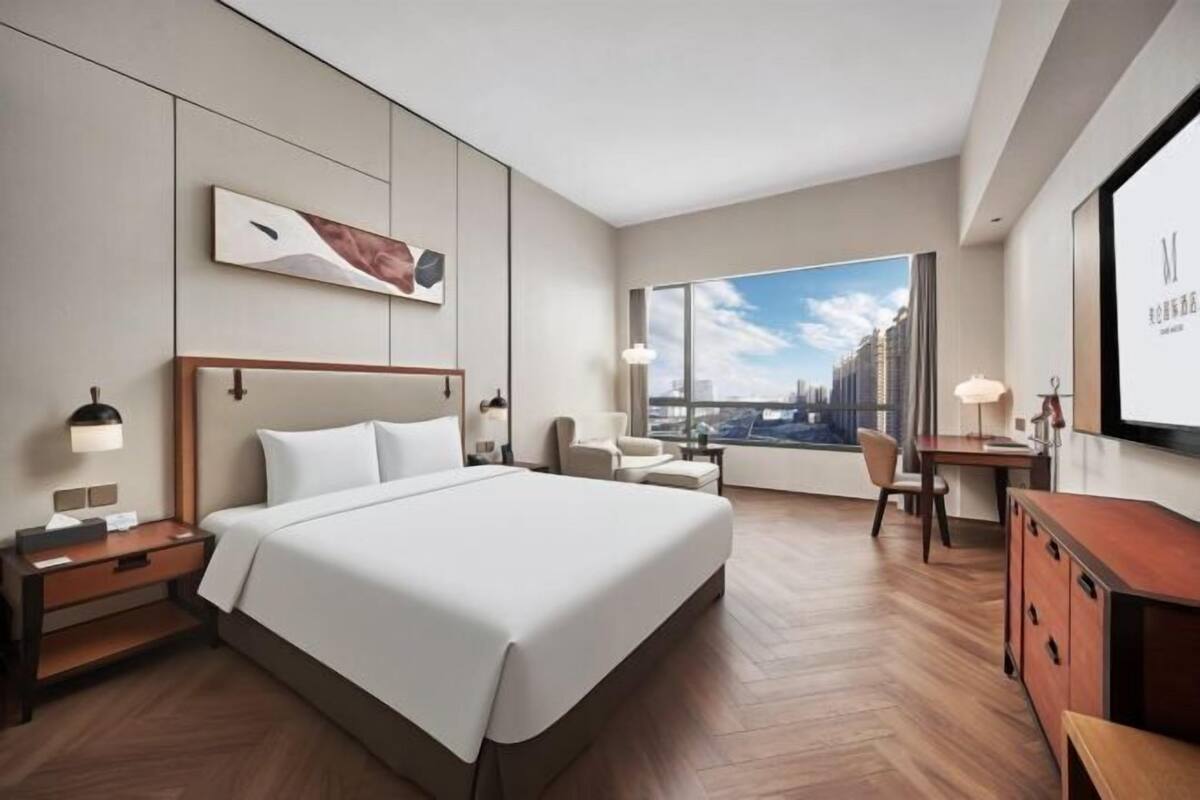 Ein modernes Hotelzimmer mit einem großen Bett, einem Schreibtisch, einem Sessel und Blick auf die Stadt durch große Fenster.