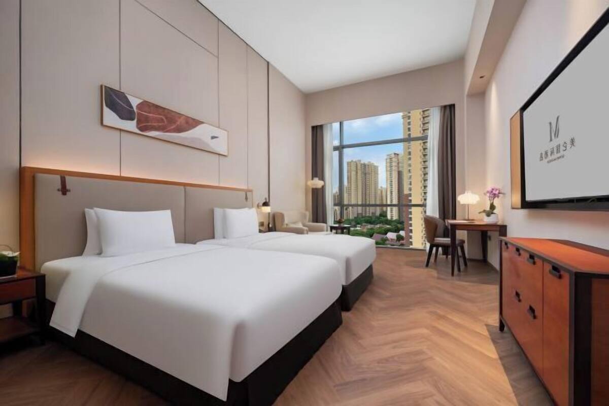 Ein Hotelzimmer mit zwei Betten, einem Schreibtisch, einem Stuhl, einem Fernseher und Blick auf hohe Gebäude.