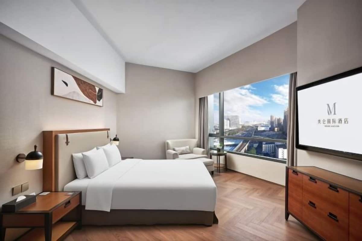 Ein modernes Hotelzimmer mit einem großen Bett, einem Schreibtisch, einem Sessel und Blick auf die Stadt durch große Fenster.