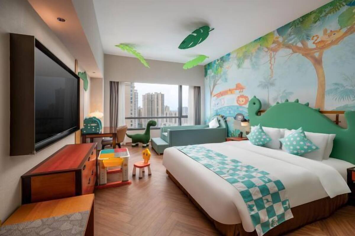 Ein Hotelzimmer mit Bett, Schreibtisch, Stuhl, Fernseher und einem Fenster mit Blick auf die Stadt.