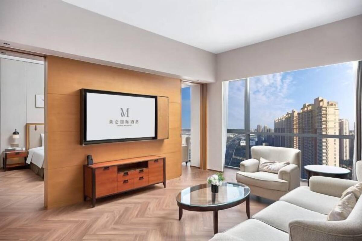 Ein modernes Hotelzimmer mit einem großen Fernseher, der an einer Holzvertäfelung angebracht ist, einer Couch, einem Couchtisch und Blick auf hohe Gebäude.